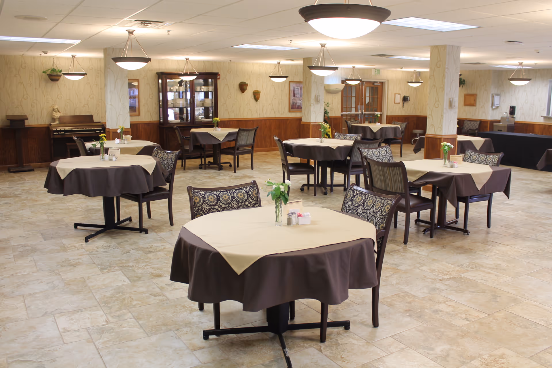 A spacious dining room with several round tables covered with beige and brown tablecloths. Each table has a small vase with flowers and salt and pepper shakers. The room has patterned chairs, tiled flooring, and warm lighting from ceiling fixtures. There is a wooden cabinet and a piano against the far wall, with light-colored wallpaper and wooden paneling on the lower half of the walls.
