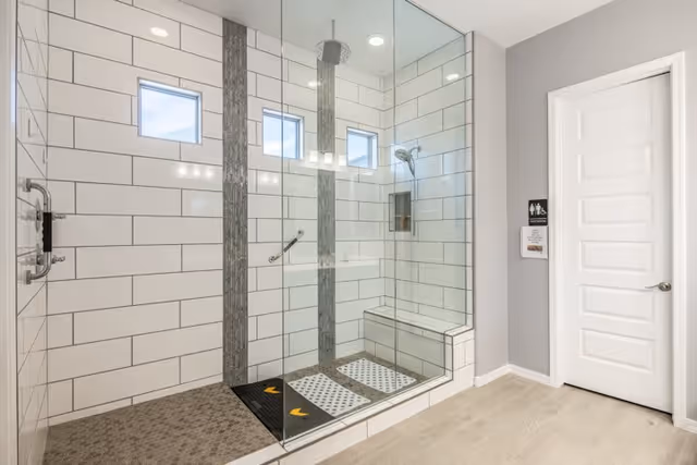 A modern, spacious shower area with glass walls and white subway tiles accented by vertical gray tile strips. The shower features a built-in bench, a rainfall showerhead, a handheld showerhead, and grab bars for safety. There are three small windows near the ceiling allowing natural light in. Adjacent to the shower is a closed white door with a sign indicating restroom facilities.