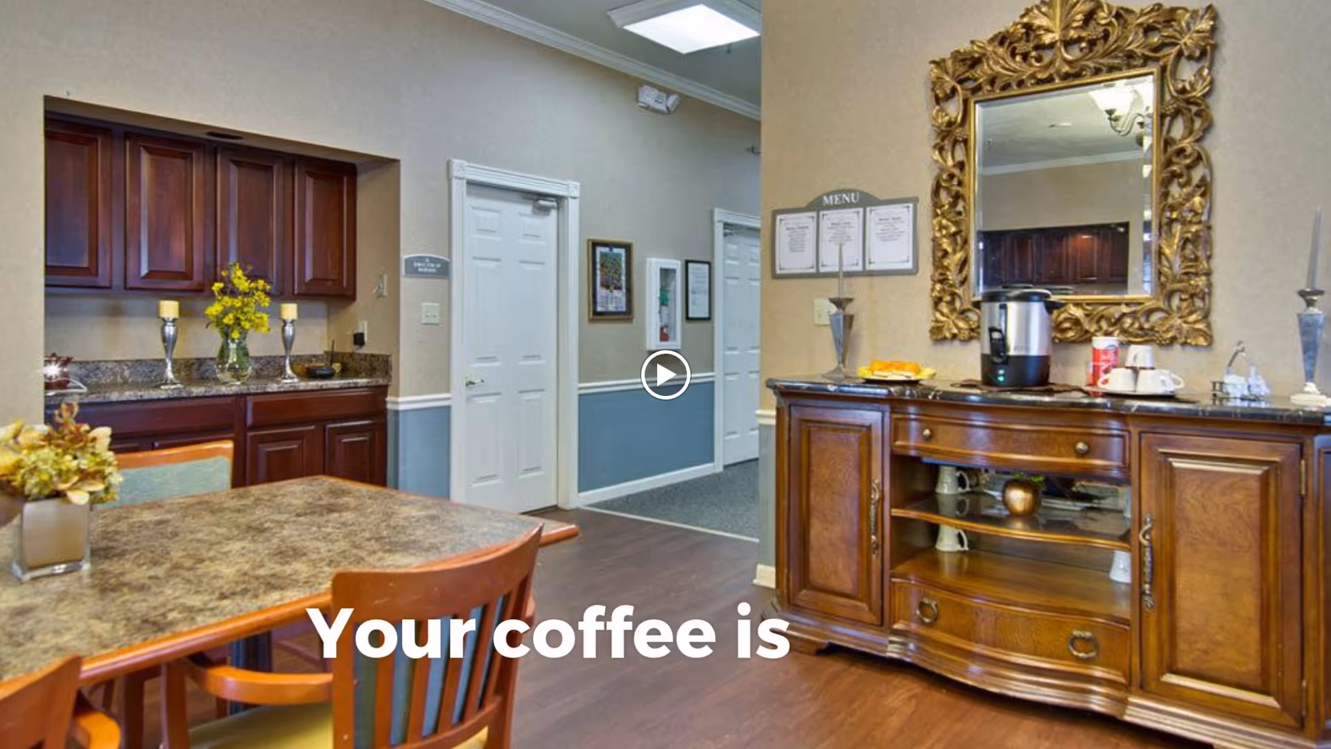 Interior view of a cozy common area with a wooden table and chairs, a sideboard with a coffee maker, cups, and a decorative mirror above it. The room has wooden cabinets, a granite countertop with candles and flowers, and a hallway with white doors in the background.