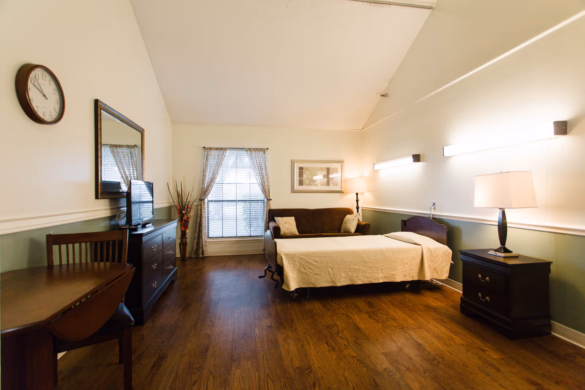 A well-lit room with wooden flooring featuring a single bed with beige bedding, a dark brown sofa behind the bed, a wooden nightstand with a lamp, a dresser with a TV and a mirror above it, a wooden table with chairs, a window with curtains, and a wall clock.