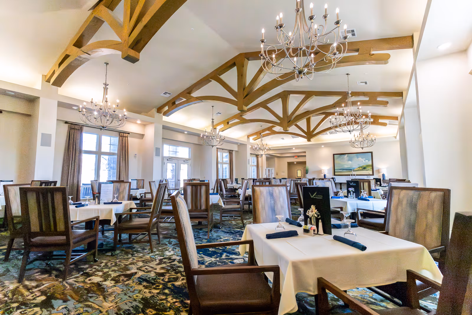 A spacious dining room with multiple tables covered in white tablecloths, each set with napkins, glasses, and menus. The room features wooden beams on the ceiling, elegant chandeliers, large windows with curtains, and a patterned carpet.