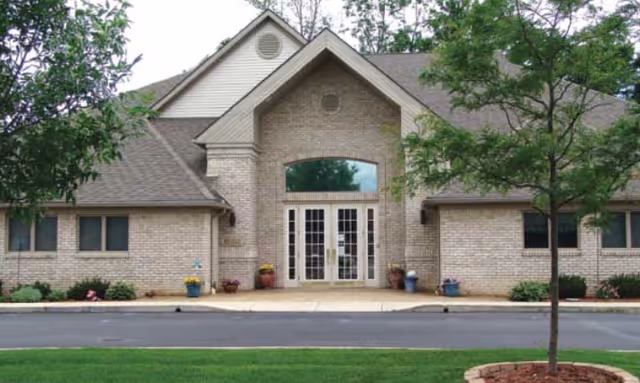 Brick building front with central double-glass doors under a gabled gable, flanked by windows, trees, and planters.