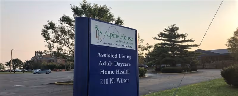 Outdoor view of a large blue sign for Alpine House of West Union, an assisted living facility, listing services including Assisted Living, Adult Daycare, and Home Health, located at 210 N. Wilson. The background shows a parking lot, trees, and a building under a clear sky at sunset.