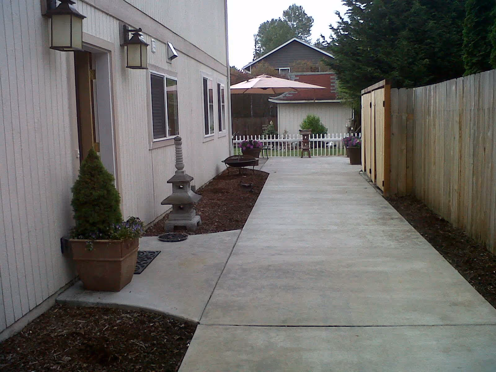 Concrete pathway alongside a beige building with potted plants, a small stone lantern, and outdoor lighting fixtures. The path leads to a backyard area with a white picket fence, a patio umbrella, and greenery including trees and shrubs.