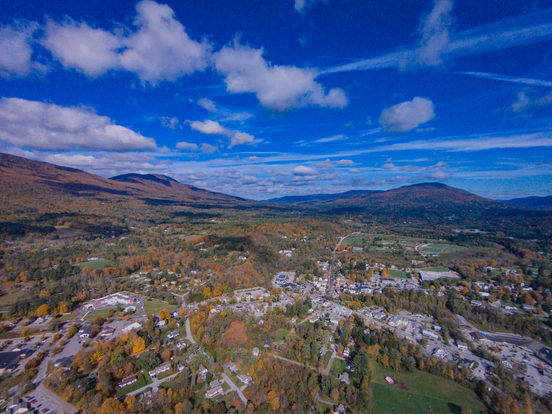 Aerial view of a small town surrounded by hills and mountains under a partly cloudy blue sky, showing residential areas, roads, and patches of trees with autumn foliage.