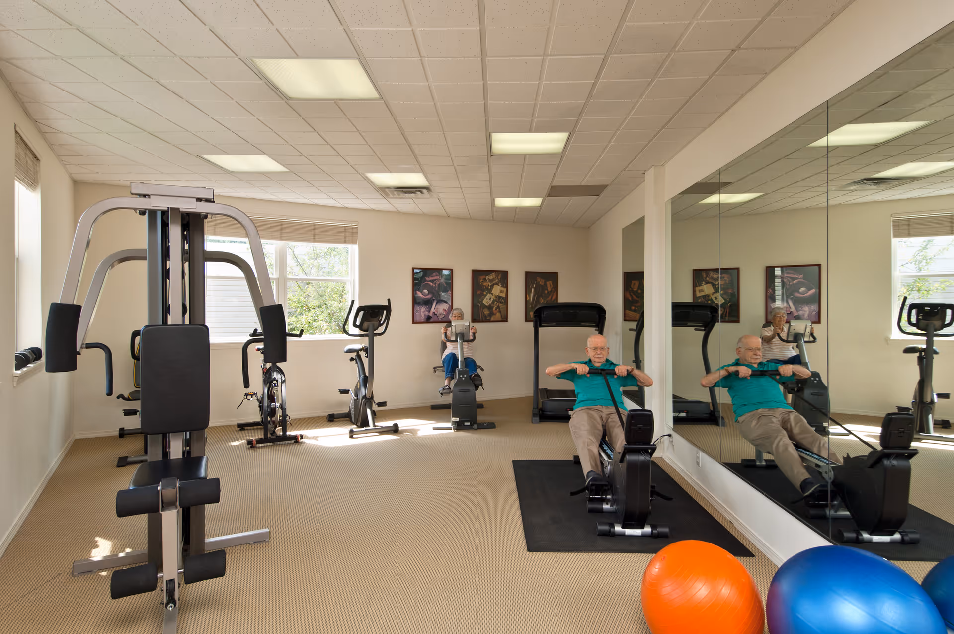 A senior fitness room with exercise equipment including a multi-gym machine, stationary bikes, and a rowing machine. Two elderly individuals are exercising, one on a rowing machine and the other on a stationary bike. The room has beige carpeting, large windows, a mirrored wall, and colorful exercise balls on the floor.