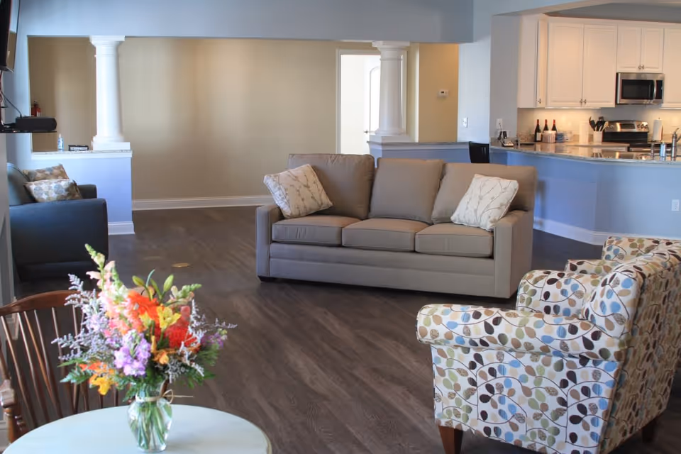 A spacious living area with a beige sofa and two patterned armchairs arranged around a small round table with a colorful flower arrangement. The room has dark wood flooring and an open layout leading to a kitchen with white cabinets and stainless steel appliances. There are two white columns separating the living area from the adjacent space.