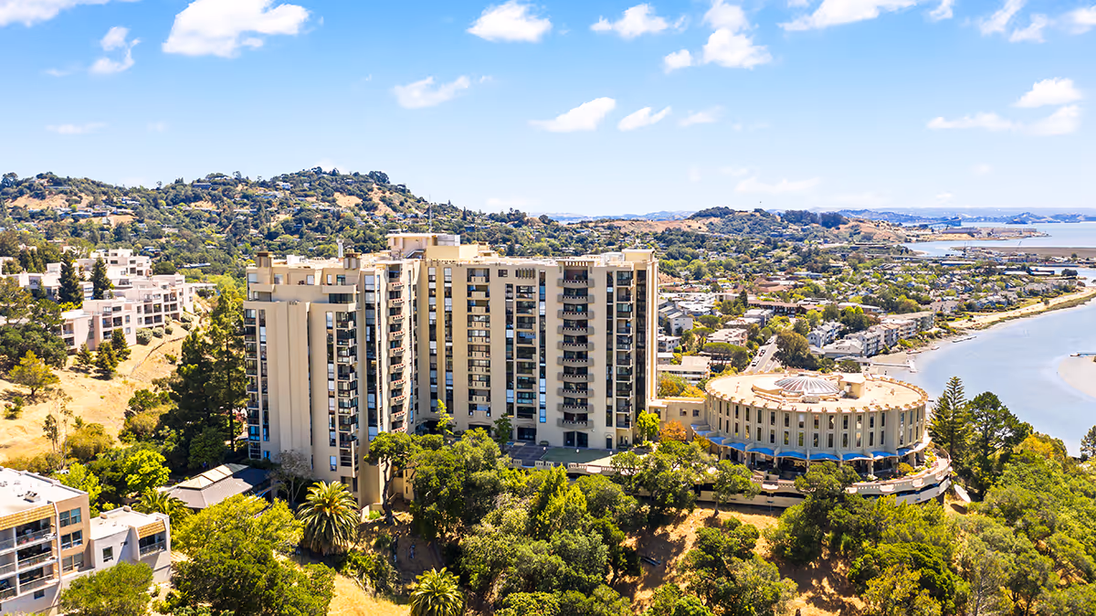 Aerial view of The Tamalpais Marin senior living facility surrounded by trees and hills, with a large multi-story building and a distinctive round building near a body of water under a blue sky with scattered clouds.