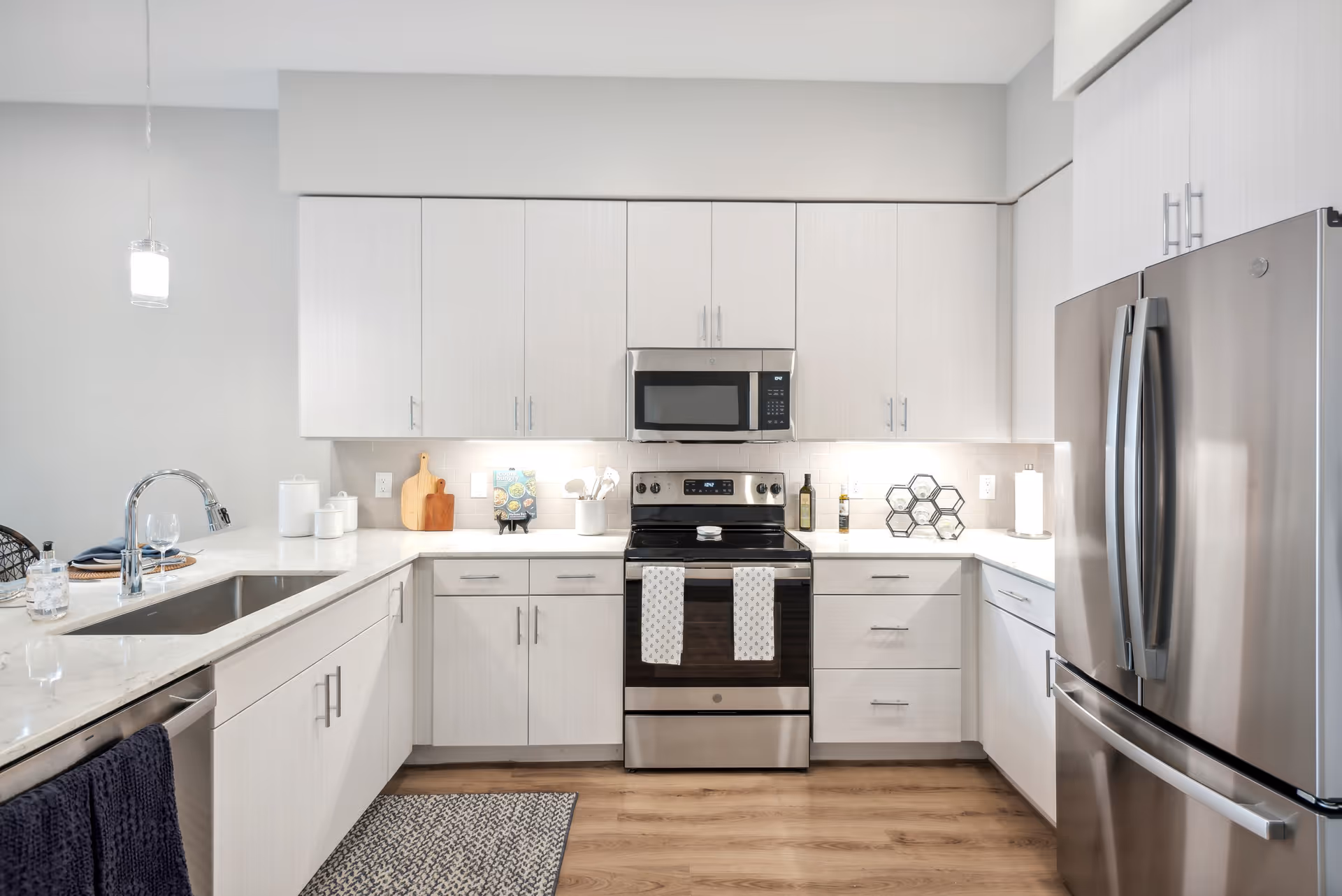 Modern kitchen with white cabinets, stainless steel appliances including a refrigerator, oven, and microwave. The kitchen features a sink with a chrome faucet, a dishwasher, and a light-colored countertop with various kitchen accessories such as cutting boards, a cookbook, and a wine rack. The floor is wooden, and there is a small rug in front of the sink.