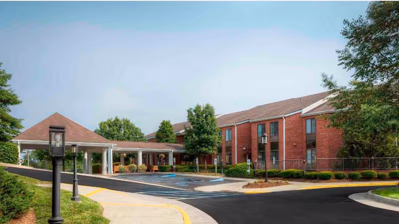 Exterior view of a red brick senior living facility with a covered entrance, surrounded by trees and landscaped bushes under a clear sky.