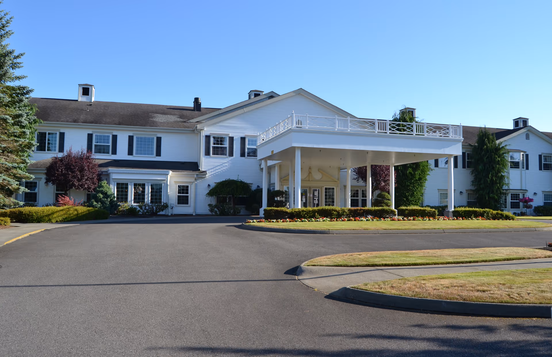 Front exterior view of a large white senior living facility building with black shutters, a covered entrance with white pillars, landscaped bushes, and a clear blue sky.