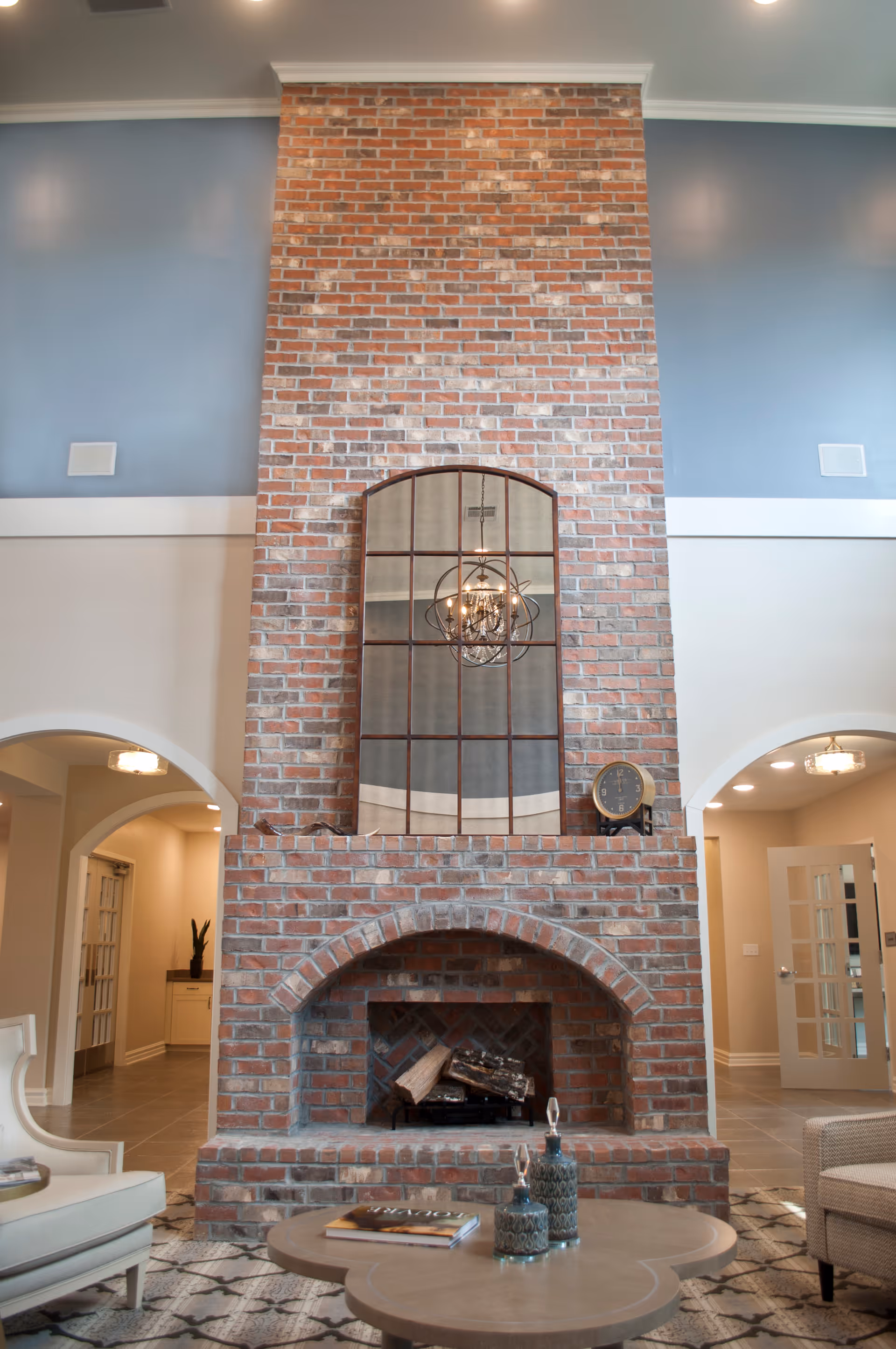 A tall brick fireplace with an arched opening in a living room. Above the fireplace is a large window with a decorative chandelier reflected in it. The room has light blue walls above white wainscoting, arched doorways on either side, and a round wooden coffee table with decorative items in front of the fireplace.