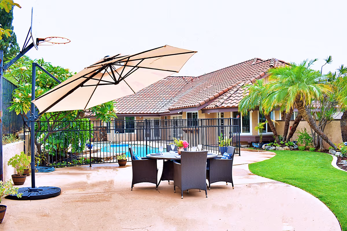 Outdoor patio area with a round table and six wicker chairs under a large beige umbrella. There is a fenced swimming pool in the background, surrounded by greenery and palm trees. The building with a tiled roof is visible behind the pool.