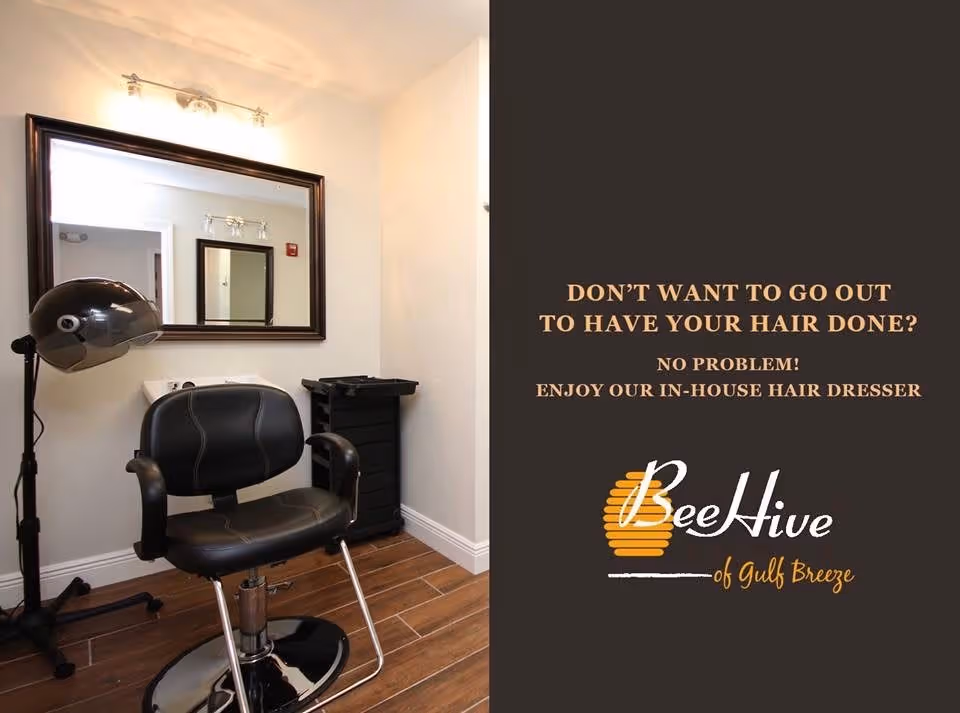 Interior view of a small hairdresser station with a black salon chair, a hair dryer on a stand, a large mirror on the wall, and a black rolling cart. The floor has wood-like tiles and the walls are painted white.