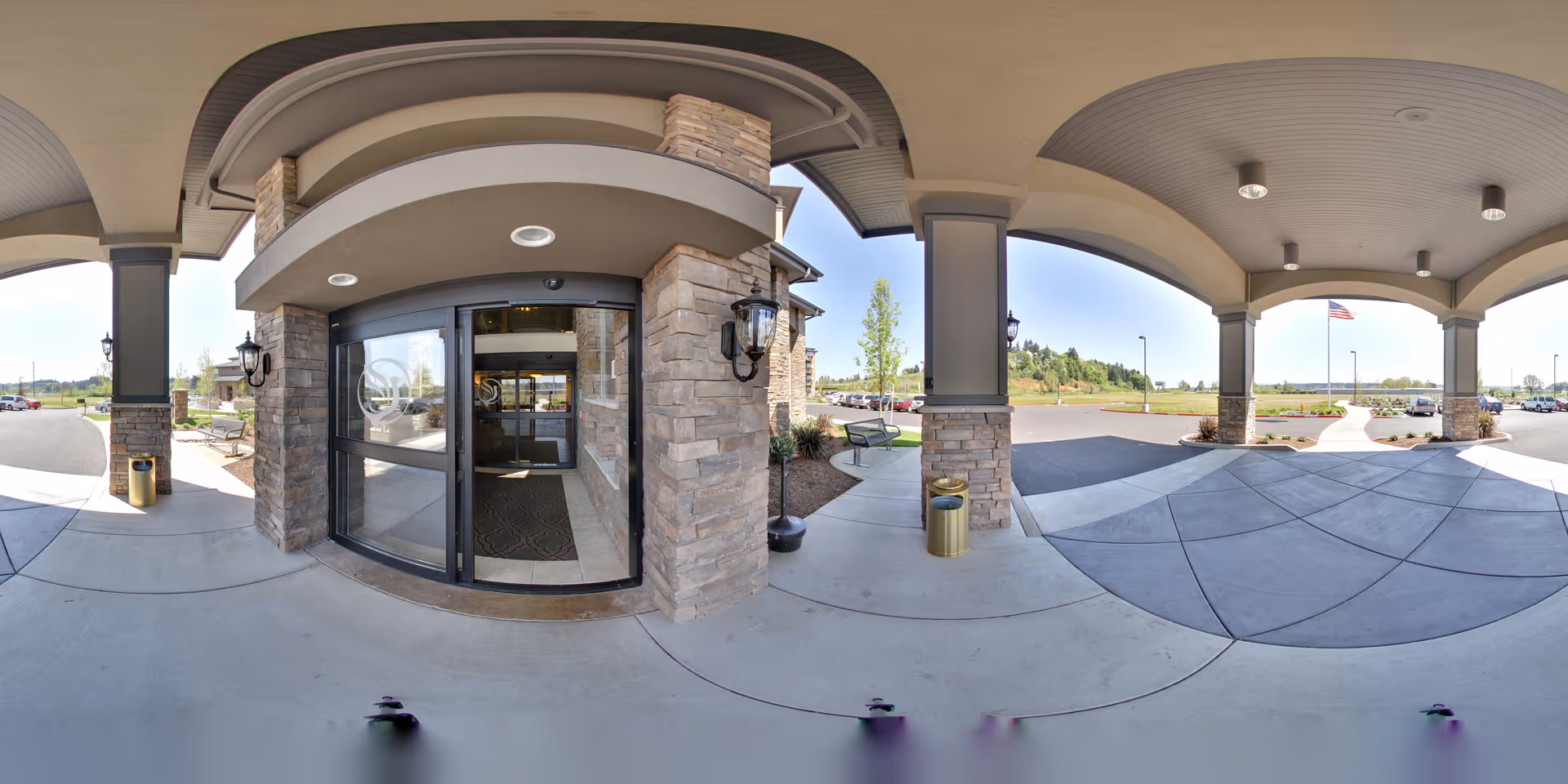 Covered entrance area of a senior living facility with stone pillars and a glass automatic sliding door. The entrance is flanked by outdoor benches, trash cans, and lantern-style wall lights. In the background, there is a parking lot, a flagpole with an American flag, and some greenery under a clear sky.