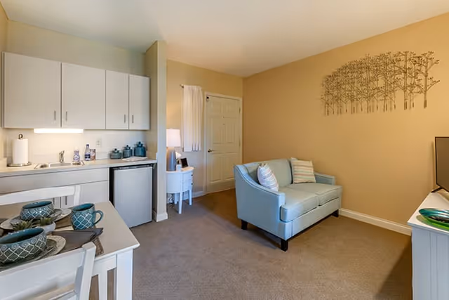 A cozy living area in a senior living facility featuring a small kitchenette with white cabinets, a mini fridge, and a countertop with various items. Adjacent to the kitchenette is a dining table set with plates, bowls, and mugs. The room also includes a light blue loveseat with two striped pillows, a small white side table with a lamp, and a wall decoration of tree silhouettes on a beige wall.