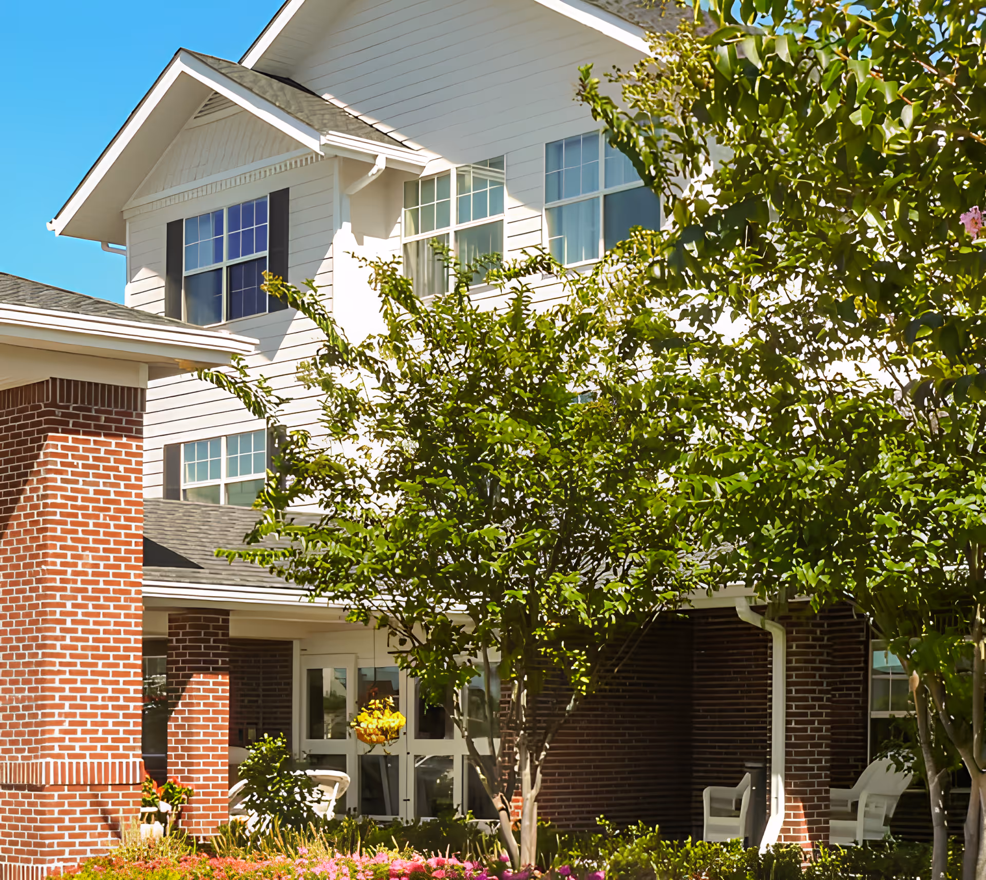 Exterior view of a two-story building with white siding and brick accents, surrounded by green trees and colorful flowers under a clear blue sky. There are white chairs on a covered porch area.