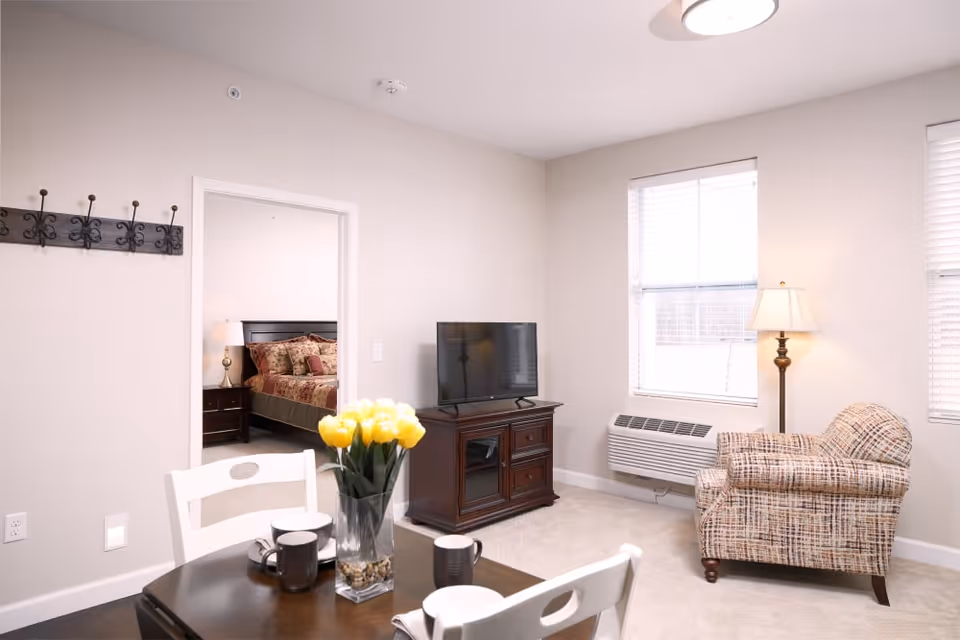 A bright living area with a patterned armchair, a wooden TV stand with a flat-screen TV, and two windows with blinds. A dining table with white chairs and a vase of yellow tulips is in the foreground. Through an open doorway, a bedroom with a bed, nightstand, and lamp is visible.