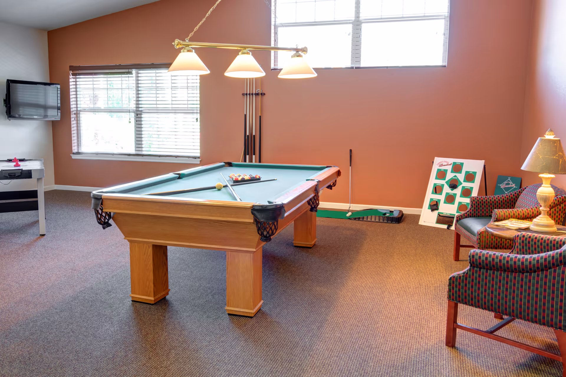 A recreational room with a pool table in the center, a wall-mounted TV on the left, a foosball table partially visible, two patterned armchairs with a lamp on a small table between them on the right, and a putting green practice set against the far wall under a window.