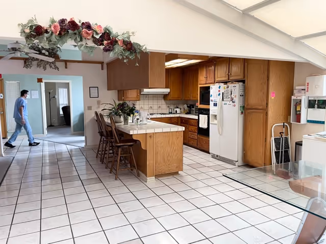 Open kitchen with tiled floor, wooden cabinets, a central island with bar stools, refrigerator and appliances, and a person walking in an adjacent room.