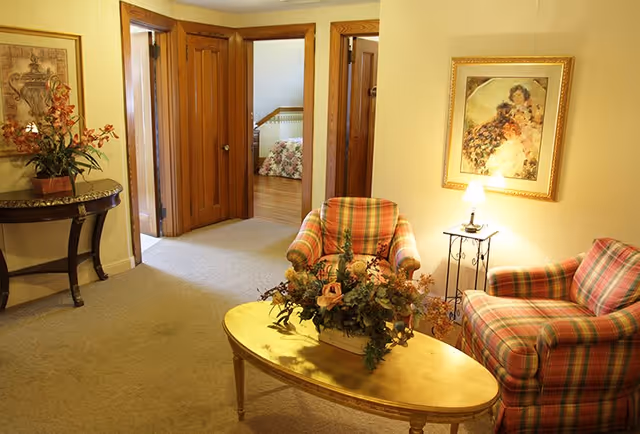 A cozy sitting area with plaid armchairs, an oval coffee table holding a floral centerpiece, and a hallway leading to adjoining rooms.