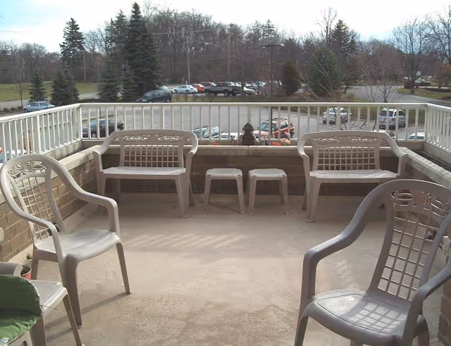 Outdoor patio area with plastic chairs and benches arranged around the perimeter. The patio has a railing and overlooks a parking lot with several cars and trees in the background.