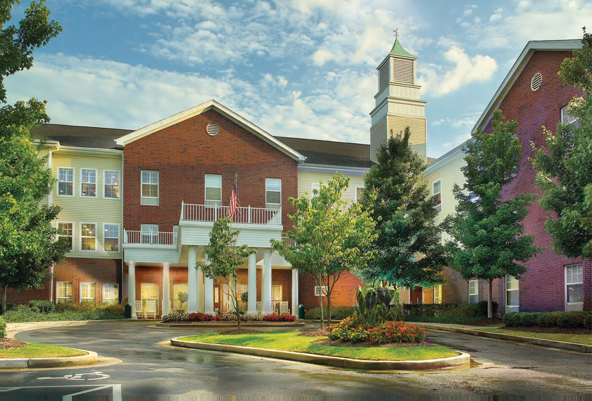 Exterior view of Belmont Village Senior Living Memphis, showing a large brick and siding building with multiple windows, a white columned entrance, an American flag, and a small tower with a green roof. The foreground includes a circular driveway, landscaped greenery, and trees under a partly cloudy sky.