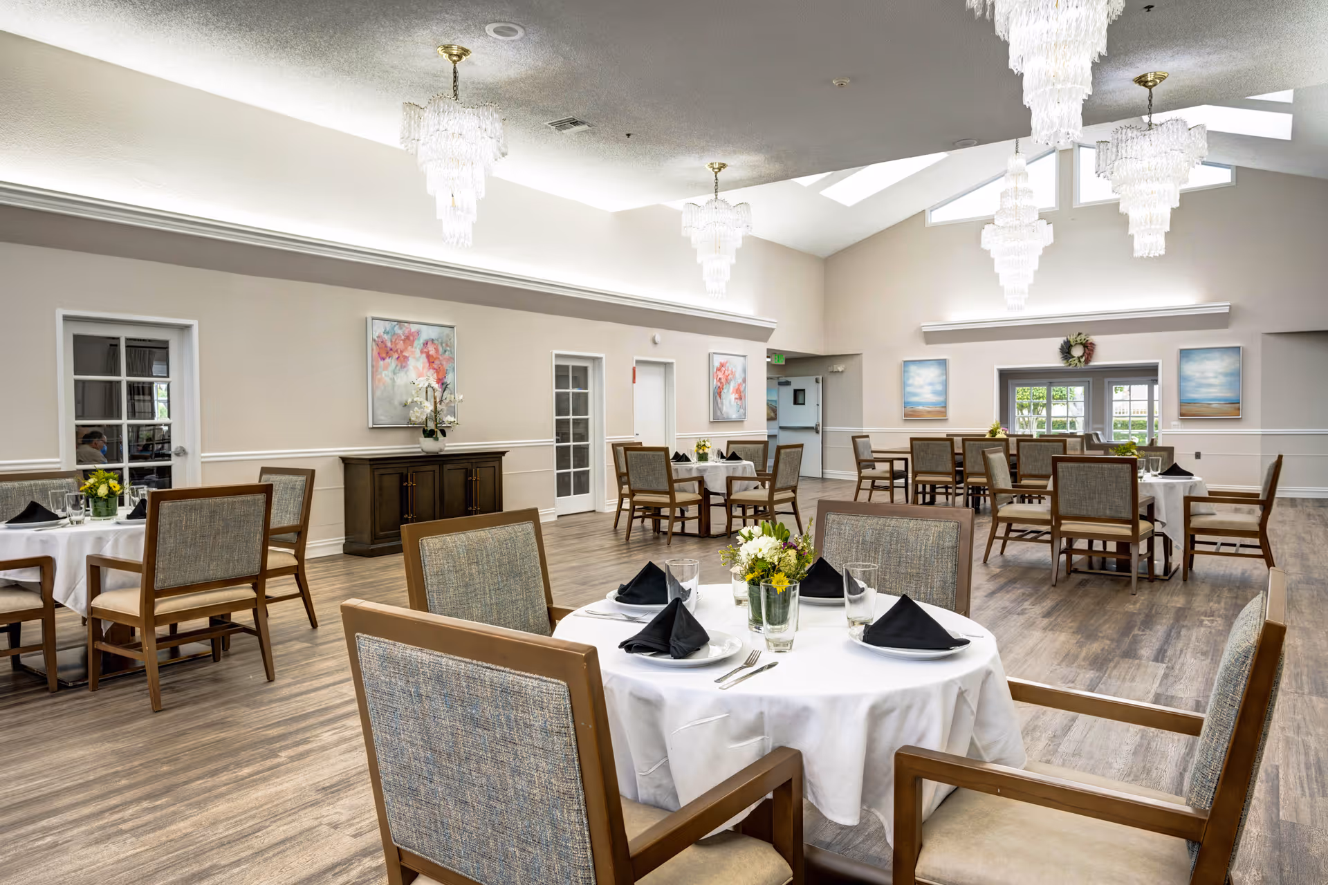 Spacious dining room with round tables set with white linens and black napkins, wooden chairs, and chandeliers under a vaulted ceiling.