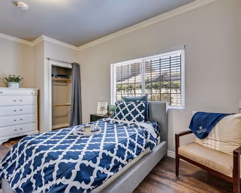 A bright bedroom featuring a single bed with a blue and white patterned comforter and matching pillow. Next to the bed is a wooden chair with a blue blanket draped over it. A white dresser with a small plant on top is positioned near an open closet with a curtain. A window with blinds lets in natural light, illuminating the room with light-colored walls and wooden flooring.