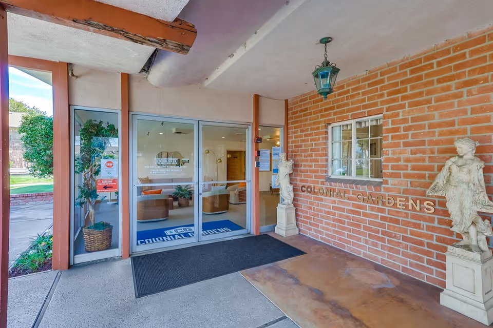Front entrance of Colonial Gardens nursing home with glass double doors, brick wall signage, statues, and an entry mat.