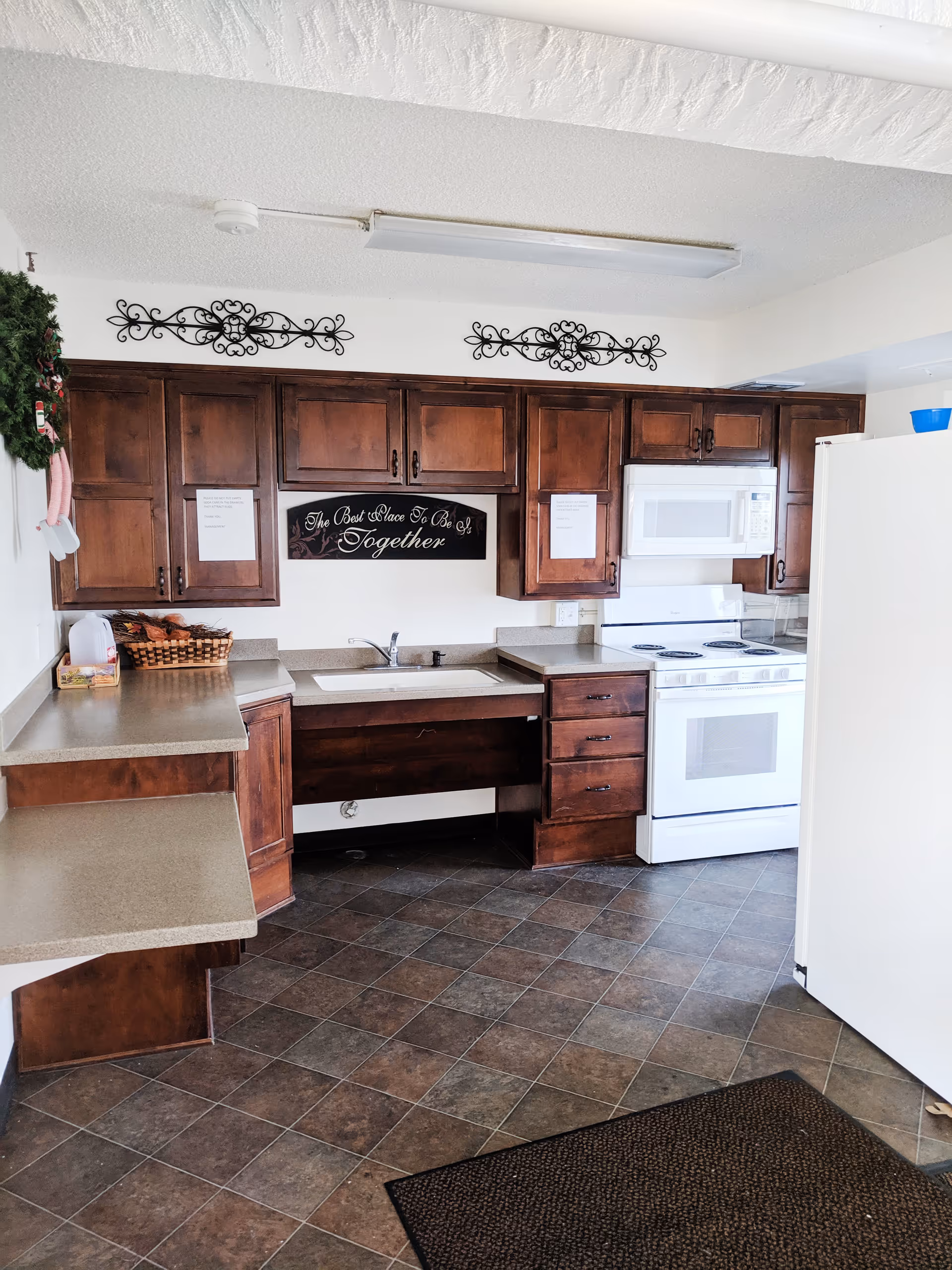A kitchen with dark wooden cabinets, a white stove with an oven, a white microwave above the stove, a white refrigerator, and a sink with a countertop. The floor is tiled with a brown and gray pattern. There is a decorative sign above the sink that reads 'The Best Place To Be Is Together' and two black ornamental wall decorations above the cabinets. A wreath is hanging on the left wall.