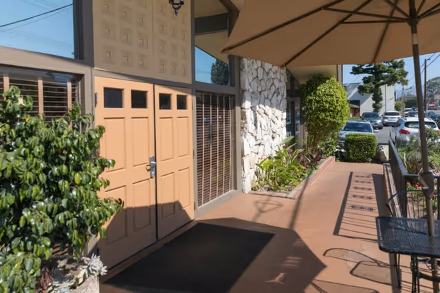 Front entrance walkway of a building with double doors, potted plants, a stone facade, outdoor umbrella and seating, and parked cars along the street.