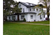 White two-story house with multiple windows, trees, and a grassy front lawn.