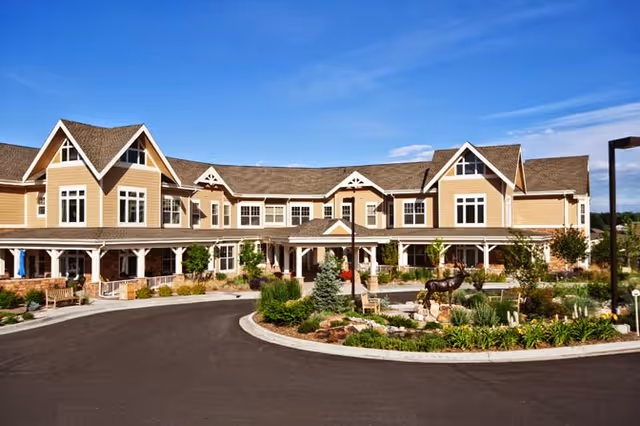 Front exterior of a two-story assisted living building with a landscaped circular driveway and sculpture under a blue sky.
