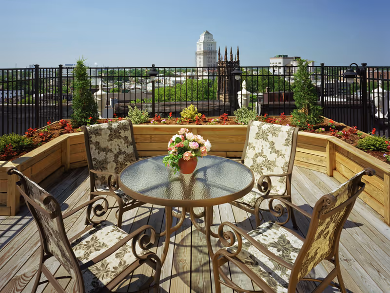 Rooftop patio with a round glass table, four patterned chairs, a potted flower centerpiece, wooden planters and a metal fence with a city skyline beyond.