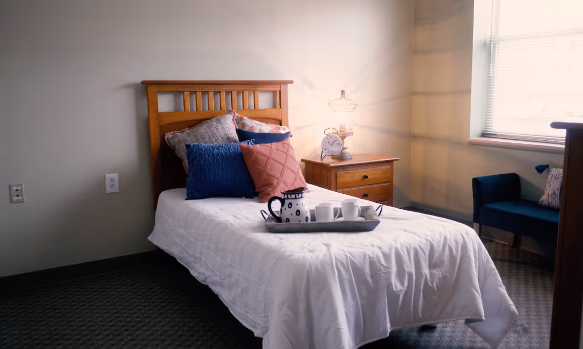 Single bed with white bedding and colorful pillows, a nightstand with a lamp and clock, and a tray on the bed in a bright bedroom.