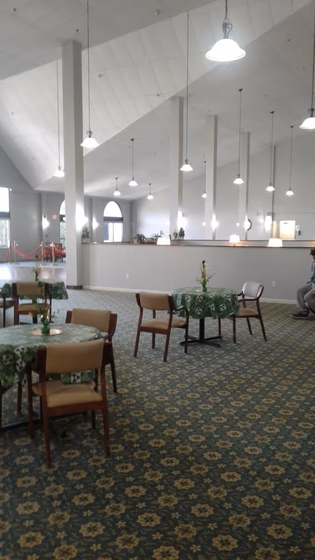 Interior view of a spacious assisted living facility common area with high ceilings and hanging pendant lights. Several small round tables covered with green patterned tablecloths are arranged on a floral carpeted floor, each surrounded by wooden chairs with beige cushions. A person is seated on the right side near the wall, and large windows and doors are visible in the background allowing natural light to enter.