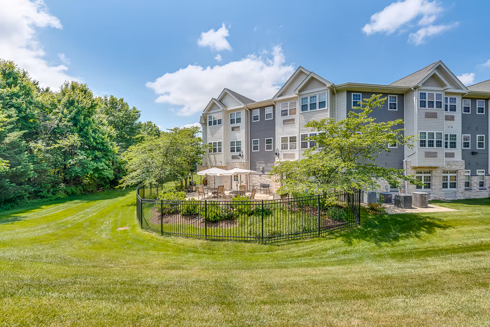 Three-story senior living building with a fenced patio, manicured lawn, and trees under a blue sky.