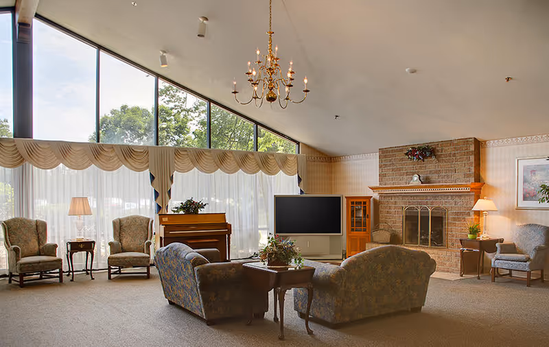 A spacious living room with large slanted windows letting in natural light. The room features a chandelier, two armchairs with a small table and lamp between them, a piano, a flat-screen TV on a stand, a brick fireplace with a decorative mantle, and two upholstered sofas around a wooden coffee table with a floral centerpiece.