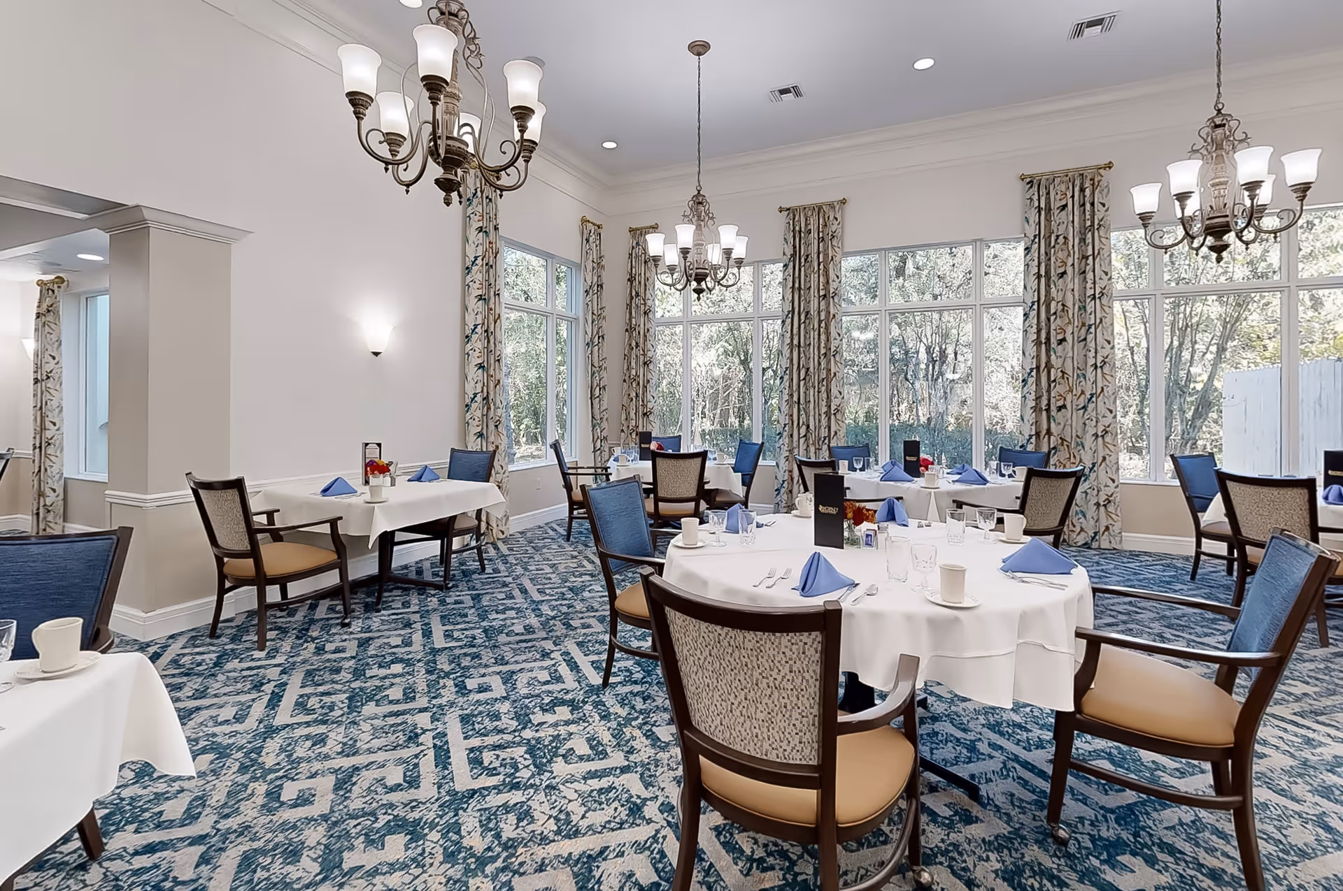 A bright dining room with multiple round and rectangular tables covered with white tablecloths, each set with blue folded napkins, cups, and silverware. The room features large windows with floral curtains, blue patterned carpet, and several ornate chandeliers hanging from the ceiling.