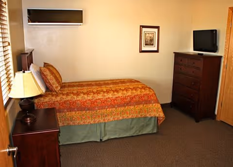Furnished bedroom with a single bed against the wall, nightstand with a lamp, dresser with a TV, and a framed picture.