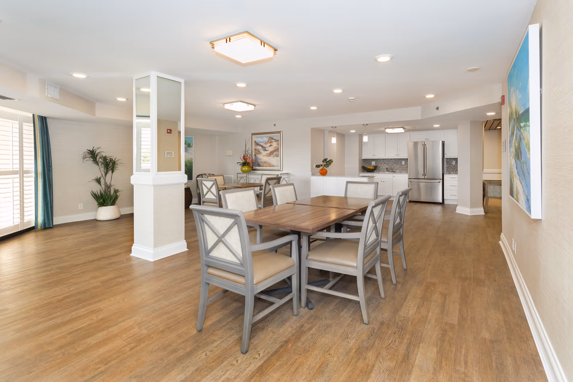 A spacious, well-lit dining area with wooden floors, several tables and chairs, a kitchen area with white cabinets and stainless steel refrigerator in the background, decorative plants, and paintings on the walls.