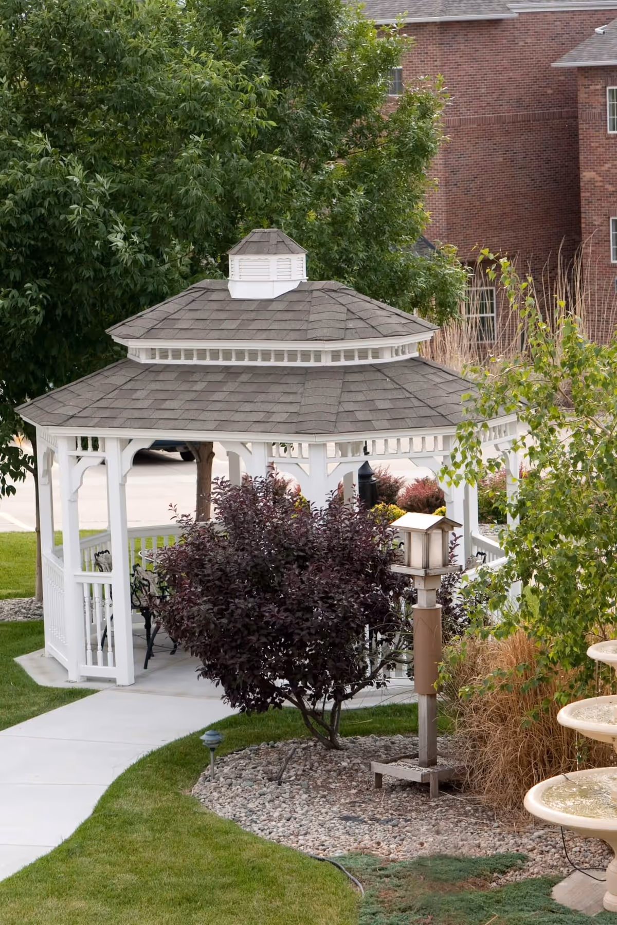 A white gazebo with a shingled roof situated in a landscaped garden area with green grass, bushes, and trees. There is a paved walkway leading to the gazebo, and a multi-tiered water fountain is partially visible on the right side. A brick building is in the background.