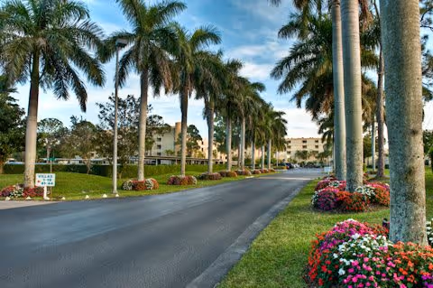 Palm-lined driveway with landscaped flower beds leading toward low-rise residential buildings under a blue sky.
