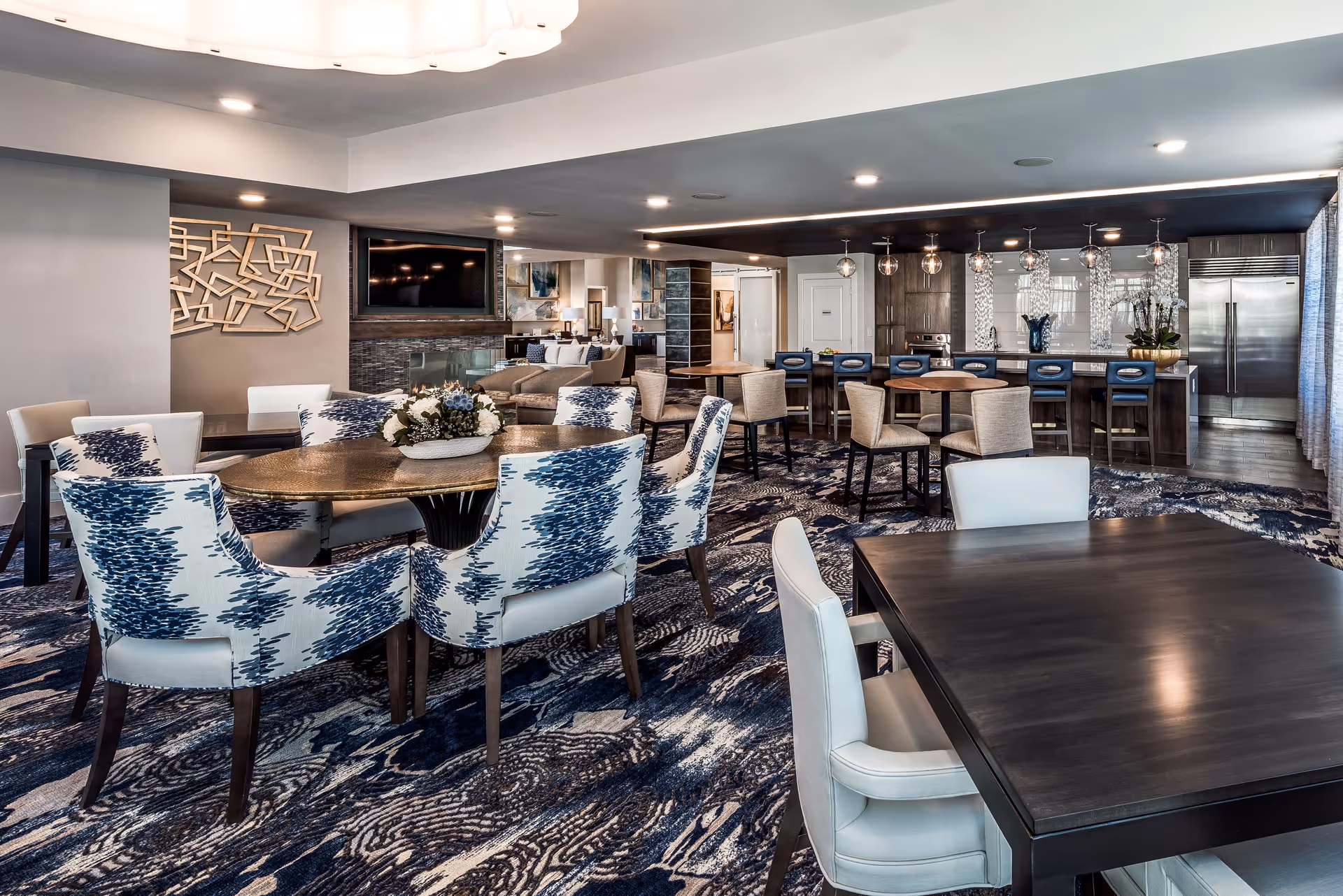 Spacious senior living common area with multiple dining tables, patterned chairs, a bar with stools, and a kitchen in the background.