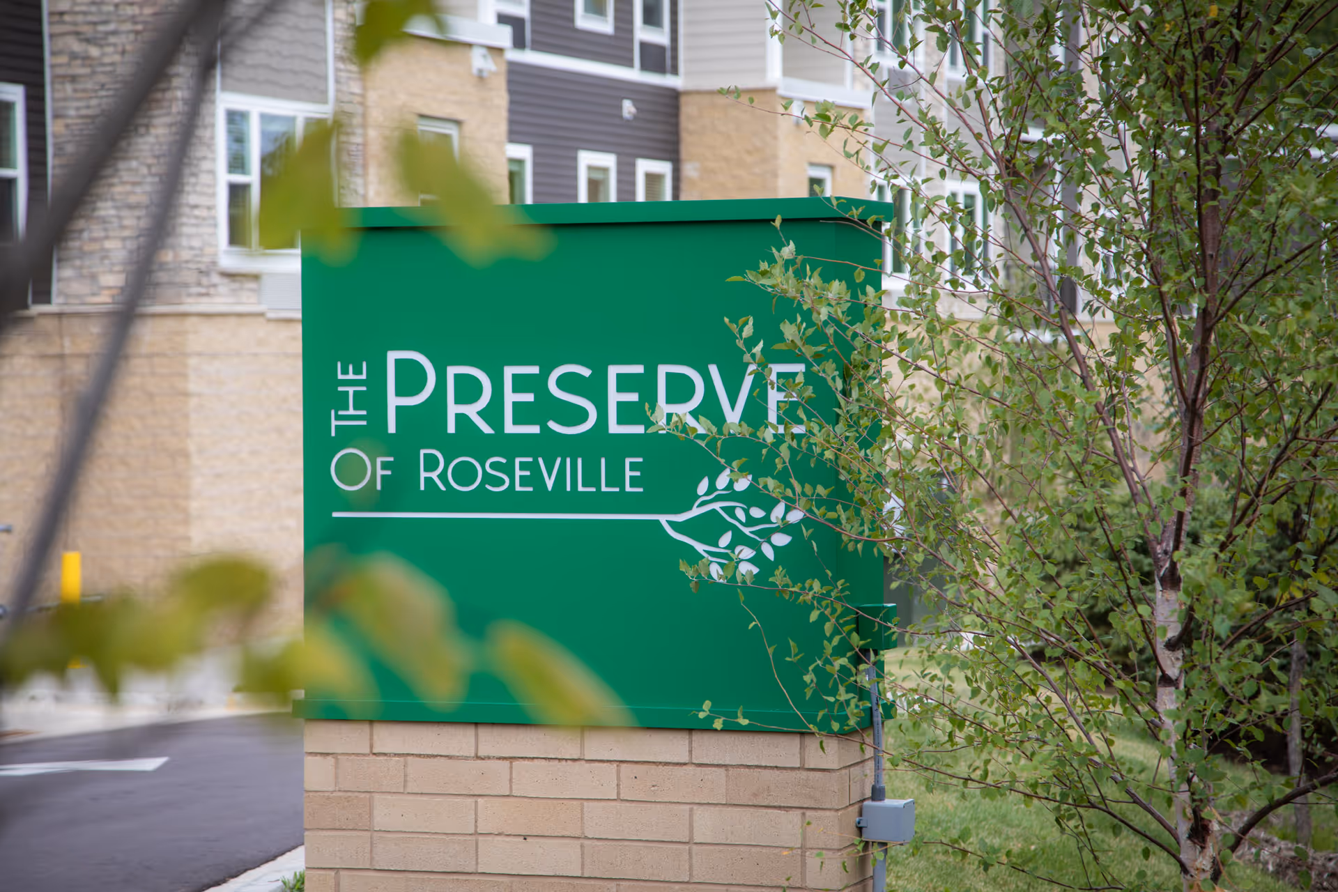 Green sign with white text reading 'The Preserve of Roseville' in front of a building with beige and gray exterior walls, partially obscured by tree branches and leaves.