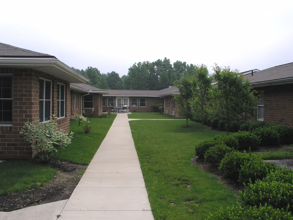 Outdoor view of a senior living facility courtyard with a concrete walkway leading through a grassy area surrounded by low bushes and small trees. The buildings are single-story with brick exteriors and multiple windows. There is a seating area with tables and chairs visible in the distance.
