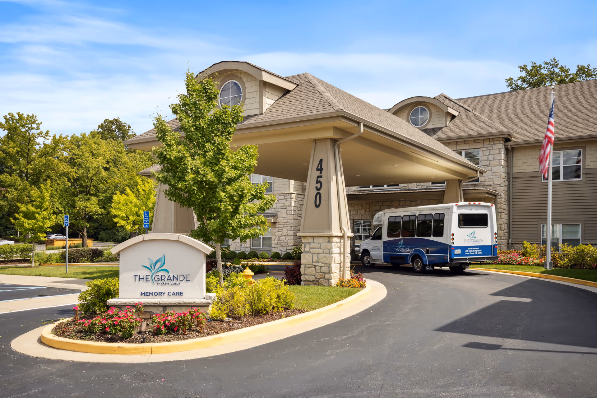 Entrance of The Grande at Creve Coeur memory care facility showing a covered drop-off area with the address number 450 on the pillar. A shuttle bus with the facility's logo is parked under the canopy. There is a landscaped area with flowers and a sign displaying the facility's name and logo. Trees and a flagpole with an American flag are visible in the background.