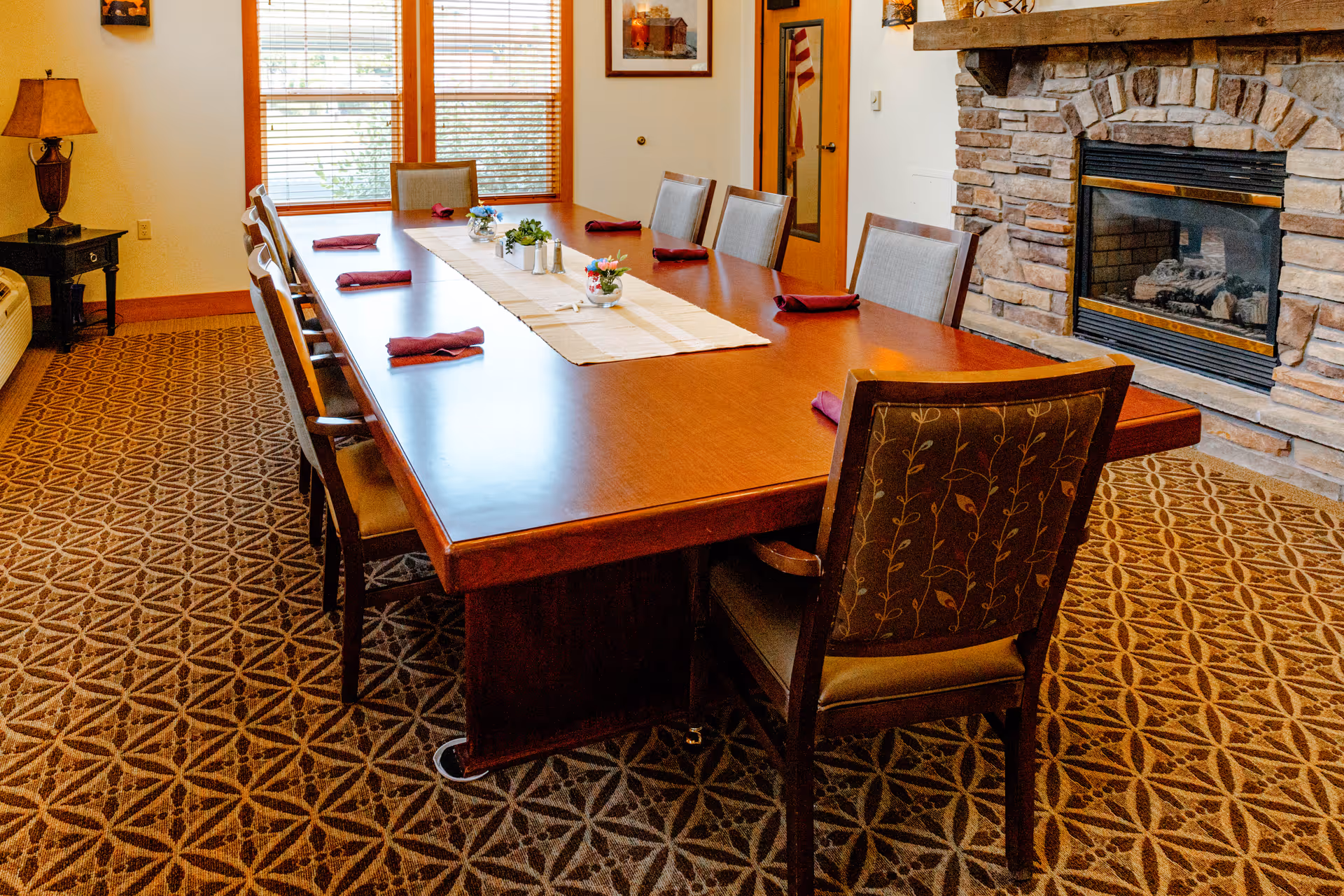 Long wooden dining table set with chairs and folded napkins beside a stone fireplace in a communal dining room.