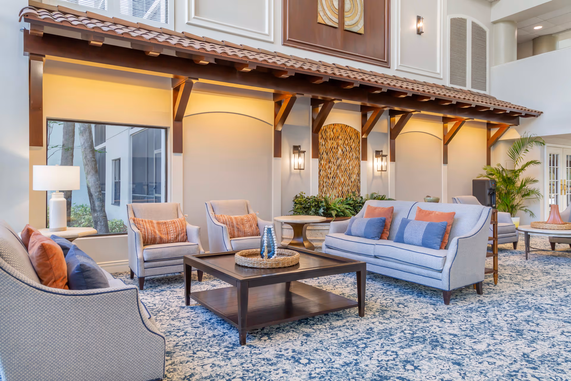 A bright and spacious living room area in a senior living facility featuring light blue upholstered sofas and armchairs with orange and blue throw pillows. A dark wooden coffee table with decorative items sits on a patterned blue and white carpet. The walls have warm lighting, decorative wall sconces, and a wooden canopy structure with a tiled roof. Large windows allow natural light to enter, and there are green plants adding a touch of nature to the space.